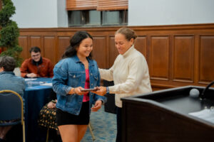 Young woman in a denim jacket receives a certificate from an older woman at a formal event in a wood-paneled room.