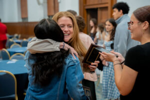 Two women embrace in a hug at an awards ceremony as another woman hands them a plaque, with people milling in the background.