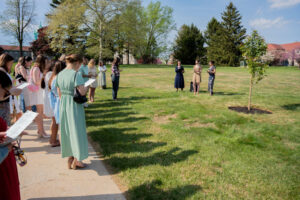 Group of attendees standing in a line along a path in a sunny park, listening to three speakers near a small tree.