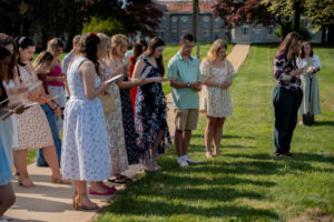Group of young adults in light dresses and casual outfits standing in a line on a sunny campus lawn, reading from papers during an outdoor ceremony.