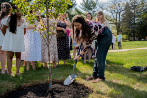 A group of young people watches as a girl digs around a newly planted tree in a sunny park setting.