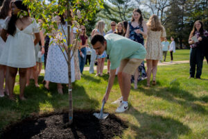 Man in a light green polo digs with a shovel as a group watches, planting a young tree in a sunny park.