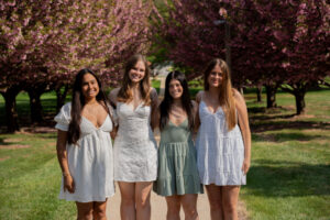 Four young women pose together in a park with blooming pink trees in the background on a sunny day, wearing light dresses.