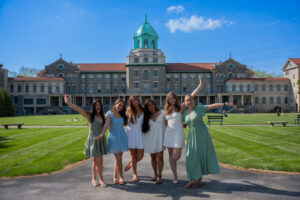 Six young women in pastel dresses pose with arms around each other on a path in front of a stately campus building with a green dome on a sunny day, green lawn surrounding them.