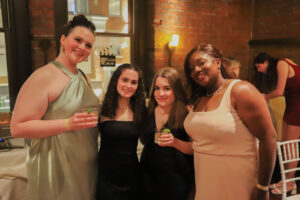 Four women posing together at a party, smiling, with drinks in hand inside a brick-walled venue.