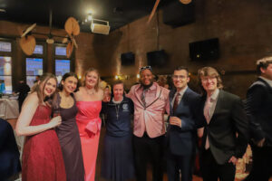 Eight people in formal attire pose together for a group photo at a brick-walled party venue with balloons and warm lighting.",