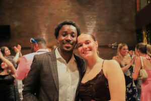 Smiling couple posing for a photo at a lively party in a brick-walled venue with guests in the background.