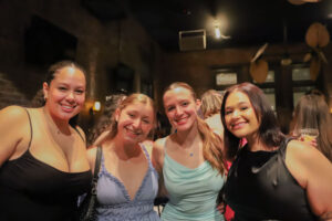 Four women smiling and posing together at a dimly lit social event in dresses (group portrait).|group photo at a party|night out with friends, four women in dresses posing for a photo