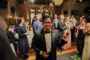 Man in a black tuxedo holds a trophy at a formal banquet, surrounded by guests in evening wear in a brick-walled venue.