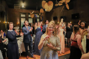 Young woman in a glittery gown smiles and holds a trophy on a dance floor as guests clap around her at a formal party.