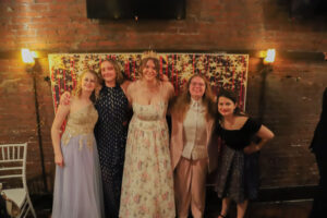 Five young women in formal dresses posing together, smiling in front of a festive backdrop at a brick-walled venue.