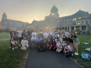 Large group of students posing for a photo on a campus path at dusk, with a historic building and string lights in the background.1) group of young people smiling outdoors on a university quad as the sun sets.2) Students gathered together for a group photo on a campus lawn in front of a historic stone building.3) Many students standing and kneeling on a path outside a grand campus building at sunset.4) Youth group photo on a university campus with warm lights and old brick architecture.