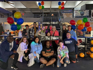Group of friends posing with a photo frame reading 'Having fun at The Palace' in a bowling alley decorated with colorful balloons and bowling balls nearby.