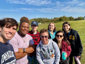 Six friends pose for a sunny outdoor selfie in a park, with a grassy field and trees in the background.