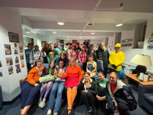 Group of about 25 people posing in a bright lounge, many smiling and wearing colorful clothes; a person in orange holds a small brown dog on their lap.