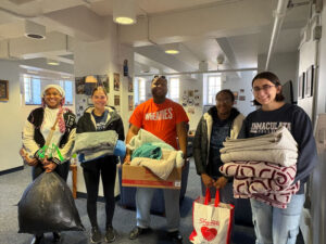 Group of five volunteers indoors, smiling and holding blankets and donation items for a charity drive.