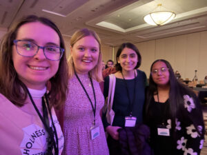 Four smiling young women wearing conference badges pose for a selfie in a hotel conference room with chandeliers in the background.