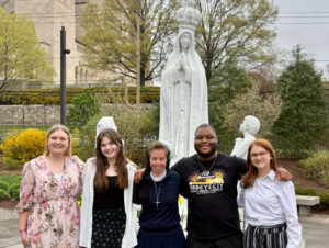 Five people pose arm-in-arm in front of a large statue of the Virgin Mary outdoors in a garden setting.