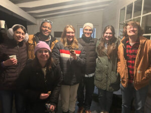 Seven young adults indoors, standing in a line in winter coats, smiling at the camera; two hold cups and one wears a pink beanie.
