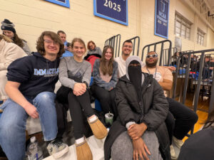 Six students sit on gym bleachers, smiling at the camera, with school banners visible on the wall in the background.