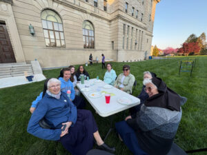 Group of nuns and volunteers seated around a white folding table on a grassy lawn, eating and chatting outside a large stone building on a sunny day.