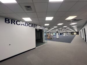 LibraryTerrace_After_2026-06-25_122 - Immaculata University Interior view of a large open space with gray carpet, white walls, and a grid ceiling with recessed lighting. A glass-enclosed room labeled "BROADCAST STUDIO" is on the left, while support columns, open walkways, and a few pieces of construction equipment are visible in the distance.