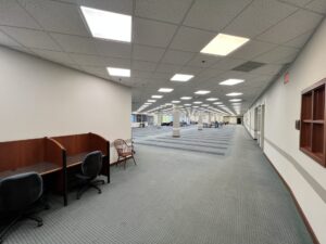 LibraryTerrace_Before_2026-04-14_40 - Immaculata University A large indoor open space with gray carpet, white walls, and a grid ceiling with recessed lighting. Two wooden study carrels with office chairs are in the foreground, while tables, chairs, support columns, and windows are visible in the distance.