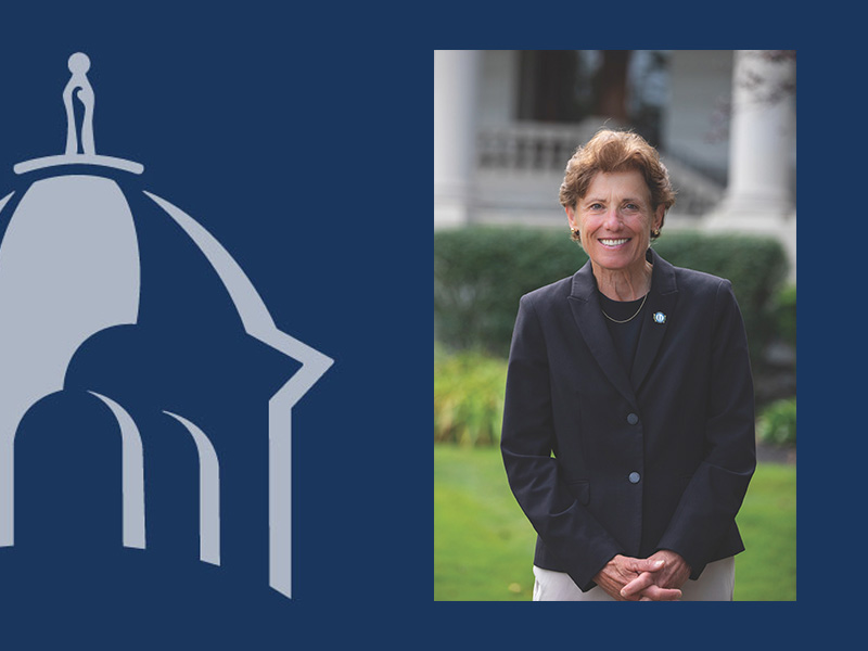 Portrait of Immaculata University President Barbara Lettiere standing outdoors, smiling at the camera. She is wearing a navy blazer over a dark top with a university lapel pin. A blue graphic featuring the university's dome logo appears beside the portrait.