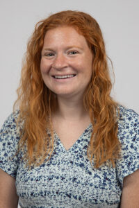 Foley_Kristen - Immaculata University Woman with long curly red hair and a floral blue blouse smiles at the camera against a gray background.