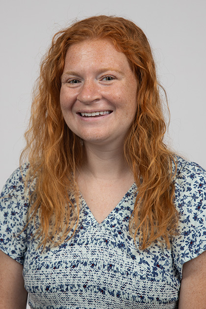 Woman with long curly red hair and a floral blue blouse smiles at the camera against a gray background.
