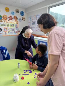 A Catholic nun in a dark habit leans over a green table inside a classroom in Peru decorated with planet cutouts, pointing toward paper crafts held by a seated boy wearing a blue sweatshirt. A young person in a light pink shirt stands beside them, watching the activity.