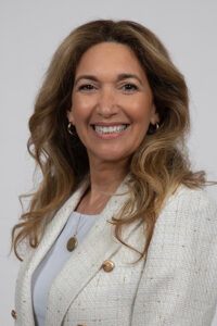 Hengst_Jacquellyne - Immaculata University Smiling woman with wavy light brown hair, wearing a white blazer and light blouse, gold earrings and necklace, posed against a neutral gray background.