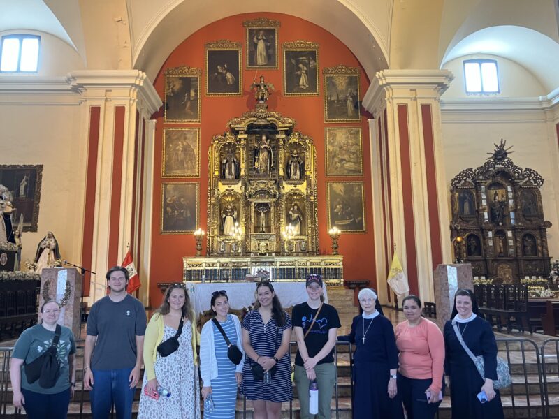 IMG_002176 - Immaculata University A group of nine people, including two Catholic nuns in habits, stand in a line facing forward in front of a metal railing inside an ornate church sanctuary. Behind them stands an elaborate, multi-tiered golden altarpiece framed by framed religious artwork set against a bright red wall, flanked by grand white and red architectural columns.