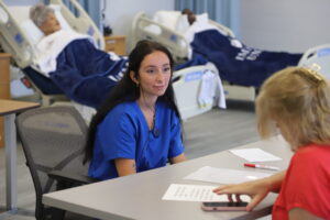 IMG_8621 - Immaculata University A student in blue medical scrubs sits at a table speaking with a faculty member during the oral exam portion of a simulation in a clinical simulation room, with two hospital beds and patient mannequins visible in the background.