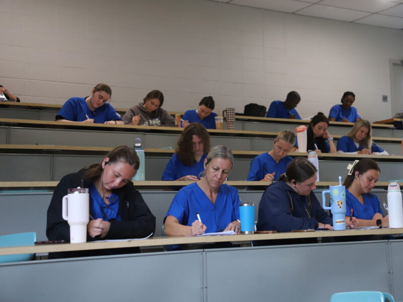 Nursing students in blue scrubs taking notes in a tiered classroom with water bottles on desks and headphones on some students.