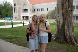 Two smiling young women with backpacks posing together on a university campus in front of a stone library building, giving thumbs up.