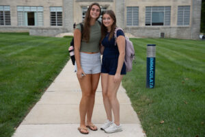 Two smiling young women with backpacks stand on a campus sidewalk in front of a stone building, posing together.