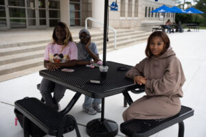 Three young people sit at a black metal picnic table outside a building, with drinks on the table and steps in the background.