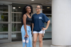 Two smiling young women pose outside a building; one wears a black tank top and light blue pants holding pink headphones, the other wears a navy T‑shirt and white shorts with glasses.