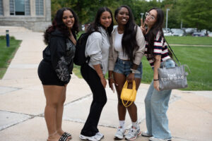 Four young women pose together outdoors on a campus path, smiling at the camera while wearing casual outfits and backpacks.