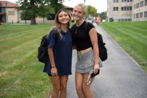 Two smiling young women with backpacks stand arm in arm on a campus path, posing for a photo in a grassy area with buildings in the background.