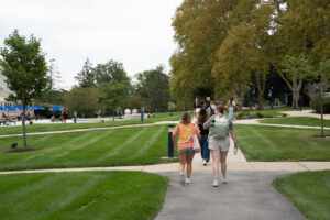 Students walk down a paved campus path through a grassy quad with trees and blue umbrellas in the background.