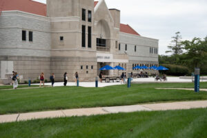 Exterior of a university library with blue umbrellas over outdoor seating and students walking along the plaza.