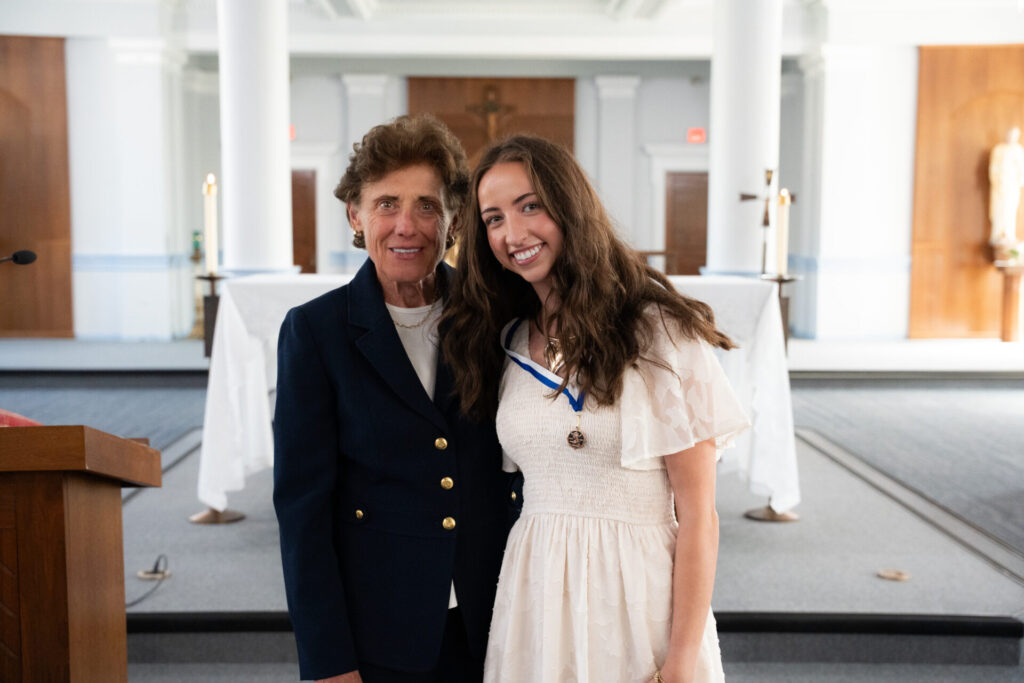 Two women smiling for a photo in a chapel. The woman on the left is in a navy blazer and cream colored shirt, while the woman on the right wears a cream-colored dress and a medal on a blue and white ribbon around her neck. They stand near an altar with white columns and lit candles in the background.