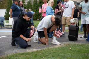 Two young men kneel on pavement, each with a small American flag, as a crowd watches at a patriotic event in a parking lot.