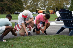 Three men kneel on a sidewalk placing small American flags in the grass at a park.
