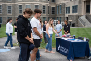 Students stand in line at an Immaculata University information table outdoors, as staff help with materials on a campus lawn. A table with brochures and American flag décor sits by the building.