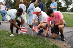Three young men kneel by a sidewalk placing small American flags in the grass during a community patriotic event.