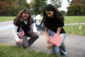 Two young women kneel on a park path, placing small American flags in the grass for a patriotic display.