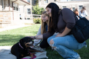 Two young women pet a small black-and-brown dog lying on a blanket on the grass.